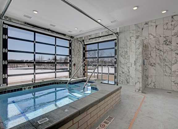 A modern indoor spa here at The Lafayette with a relaxing hot tub, marble tile walls, and glass garage-style doors. A walk-in shower and bright natural light create a refreshing, upscale wellness space.