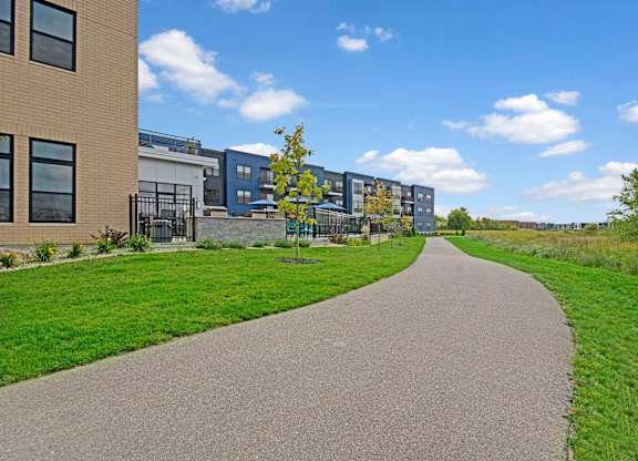 A scenic walking path here at The Lafayette with lush green landscaping and modern apartment buildings. A nearby patio area and open green space create a peaceful, inviting outdoor setting.