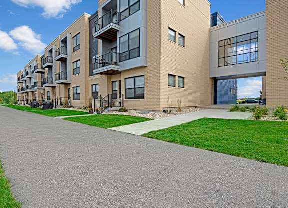 A modern exterior here at The Lafayette with private balconies, large windows, and light brick siding. A paved walking path and landscaped greenery create a clean, welcoming community setting.