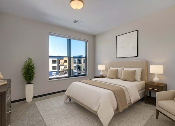 A peaceful bedroom here at The Lafayette featuring soft neutral bedding, matching nightstands with lamps, a light area rug, a cozy accent chair, and a large window bringing in bright, refreshing natural light.