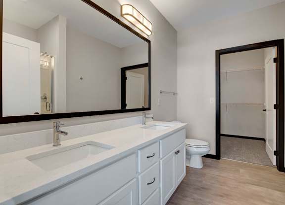 A bright bathroom here at The Lafayette featuring a wide double-sink vanity with white cabinetry, a large mirror with modern lighting, and an open doorway leading to a spacious walk-in closet.
