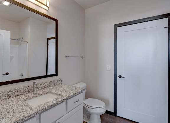 A clean bathroom here at The Lafayette featuring a bright white vanity with speckled granite countertop, a large framed mirror with modern lighting, and a sleek white door creating a fresh, inviting look.