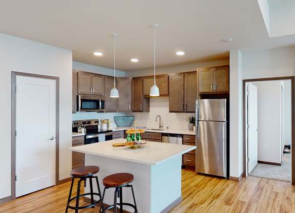 A modern kitchen here at Van Buren East with stainless steel appliances, wood-style flooring, and warm brown cabinets. A center island with bar seating and pendant lighting creates a bright, stylish, and inviting space.