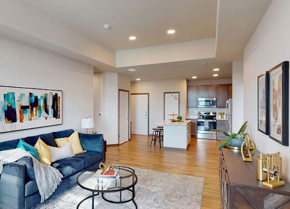A stylish living room here at Van Buren East with wood-style flooring and modern furnishings. A cozy sofa and open layout flow into a kitchen with stainless steel appliances and a center island, creating a bright, inviting space.