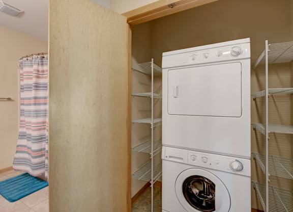 A laundry room here at Van Buren Place featuring a stacked washer and dryer with built-in shelving, creating a convenient, organized, and practical space with easy access near the bathroom.