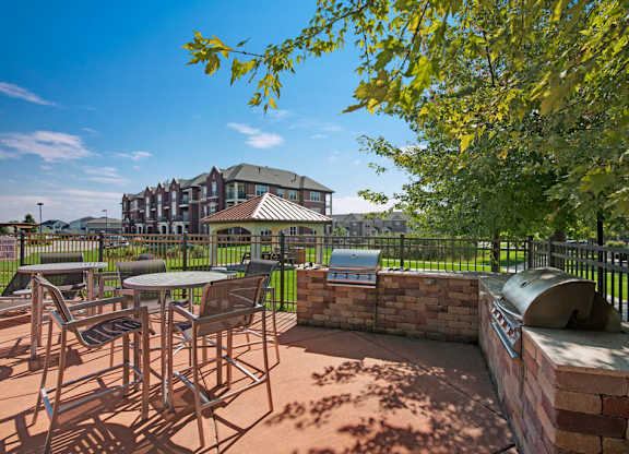 A sunny outdoor grilling patio here at Van Buren Place with built-in barbecue stations and comfortable table seating. A nearby gazebo, green lawn, and surrounding apartments create a welcoming, resort-style atmosphere.