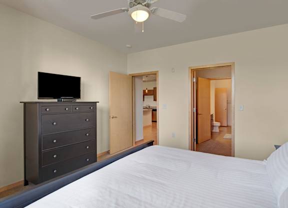 A bedroom here at Van Buren Place featuring a dark wood dresser, soft carpet flooring, an overhead ceiling fan, and an open layout with direct access to the bathroom that feels comfortable and convenient.