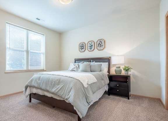 A bedroom here at Van Buren Place featuring soft carpet flooring, a dark wood bed frame, matching nightstand, and a large window that makes the space feel bright, calm, and inviting.