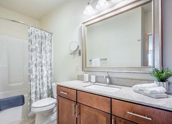 A bathroom here at Van Buren Place featuring a spacious vanity with warm wood cabinetry, large mirror, and a shower-tub combo that makes the space feel clean, bright, and comfortable.