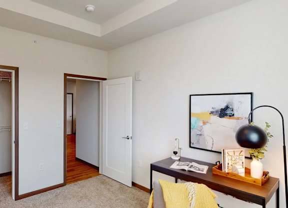 A bedroom here at Van Buren West featuring light neutral walls, an open closet with built-in shelving, soft carpet, and a dark wood desk styled with a black dome lamp and yellow accent pillow, creating a bright, functional, and welcoming space.
