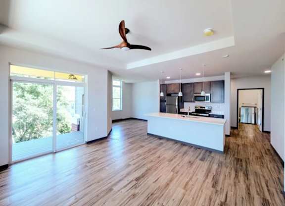 A living room here at Van Buren West featuring warm wood-style flooring, a modern kitchen island with pendant lighting, stainless steel appliances, and large sliding glass doors that fill the open layout with bright natural light.