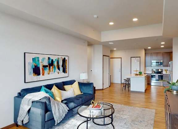 A living room here at Van Buren West featuring a deep blue sofa with yellow and teal accent pillows, round glass coffee table, light area rug, and warm wood flooring, creating a bright, open, and inviting space that flows seamlessly into the modern kitchen.