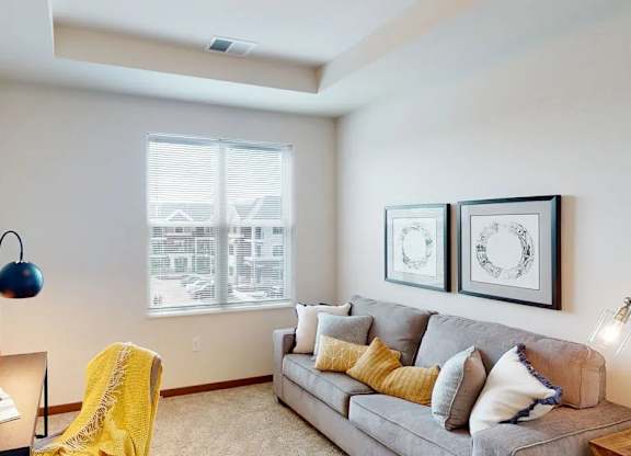 A living room here at Van Buren West featuring a light gray sofa with yellow and neutral accent pillows, soft carpet flooring, warm wood trim, and a cozy desk nook with a navy lamp and bright yellow throw for a welcoming feel.
