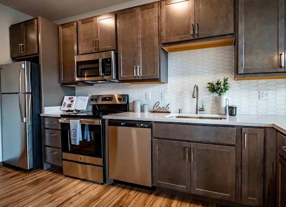 A kitchen here at Van Buren West featuring rich brown cabinets, stainless steel appliances, bright white countertops, and a crisp hexagon tile backsplash for a clean, modern, and inviting feel.