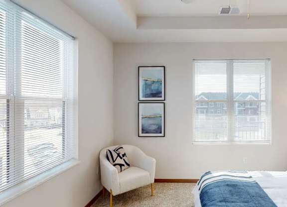 A bedroom here at Van Buren West featuring light neutral walls, large bright windows, a soft gray upholstered bed with blue accents, a wood nightstand with a purple glass lamp, and a cozy cream accent chair for a calm, inviting feel.
