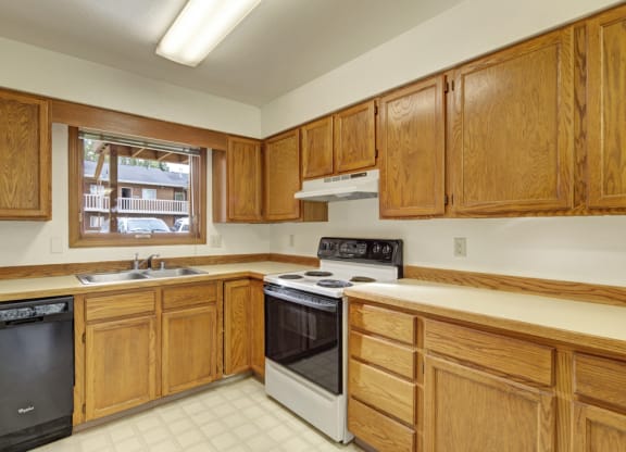 A kitchen here at Wildwood Estates Apartments with wooden cabinets, a stove, and dishwasher. A window above the sink overlooks a parking area. Bright and clean ambiance.