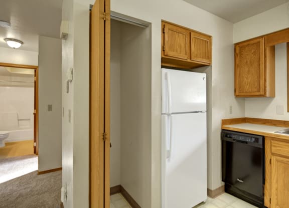 A kitchen here at Wildwood Estates Apartments with wooden cabinets, a white refrigerator, and a black dishwasher. Adjacent hallway leading to a bathroom, with a warm and clean ambiance.