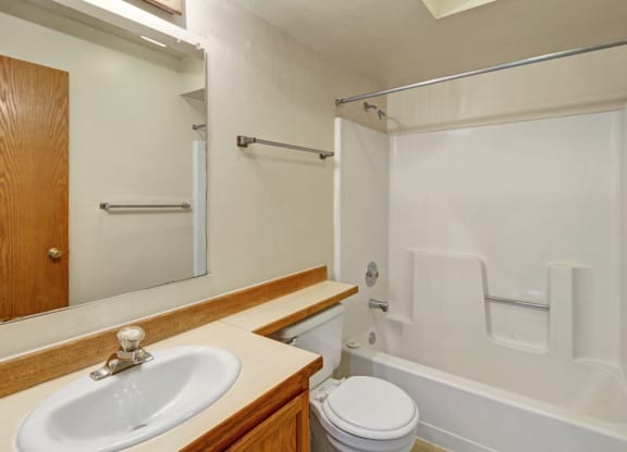 Small, tidy bathroom here at Wildwood Estates Apartments with white fixtures. A sink with wooden cabinet, toilet, and bathtub with shower inside are visible. Neutral color tones.