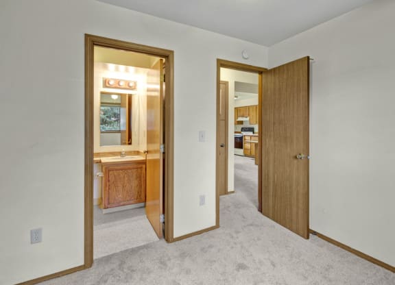 A beige carpeted room here at Wildwood Estates Apartments with two doors; one leads to a lit bathroom featuring a wooden vanity, and the other to a kitchen with wooden cabinets.
