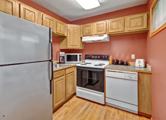 Compact kitchen here at Wildwood Estates Apartments with wooden cabinets and red walls. Features a stainless steel fridge, stove, microwave, dishwasher, toaster, jars, and wooden floor. Cozy atmosphere.