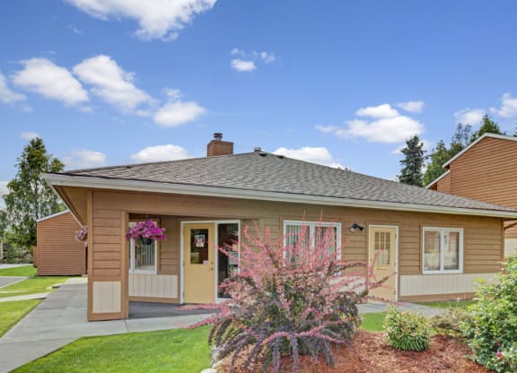 A single-story, brown wooden house here at Wildwood Estates Apartments with a grey roof sits under a bright blue sky. Pink flowers and green shrubs adorn the front, adding a welcoming touch.