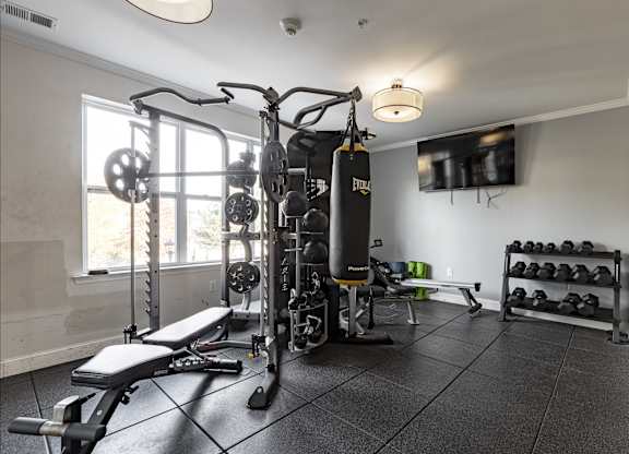 Fully equipped fitness center at Queens Gate Apartments featuring weight training equipment, a punching bag, dumbbells, and a flat-screen TV for an enhanced workout experience.