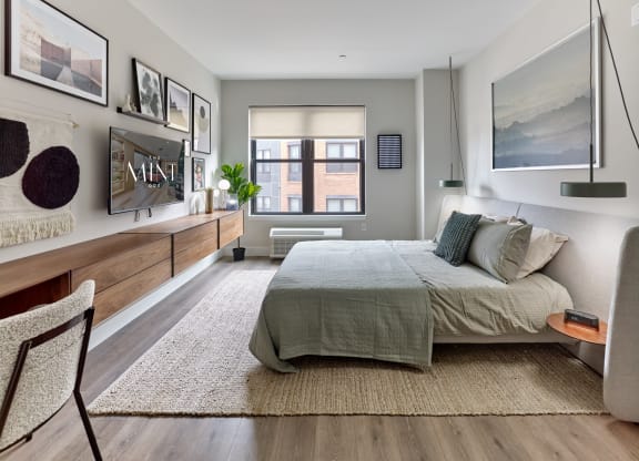 Elegant bedroom in a studio apartment at The Mint Rahway, showcasing a cozy bed, stylish decor, and a large window providing ample natural light.