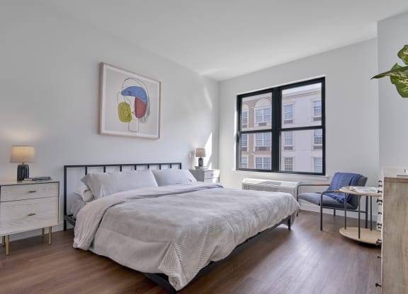 Cozy bedroom at The Mint Rahway featuring a large window, modern artwork, and comfortable furnishings, perfect for studio apartments close to the Rahway Train Station.