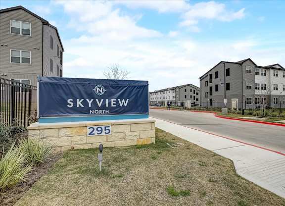 Landscaped community entrance with stone monument feature, sidewalk, and internal roadway leading to apartment homes.