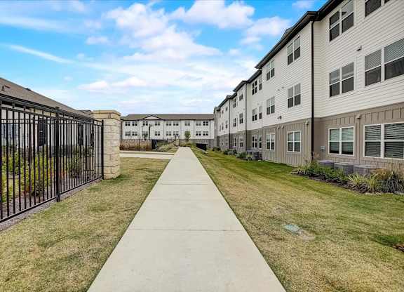 Wide sidewalk between apartment buildings, bordered by lawns, shrubs, and a secure perimeter fence.