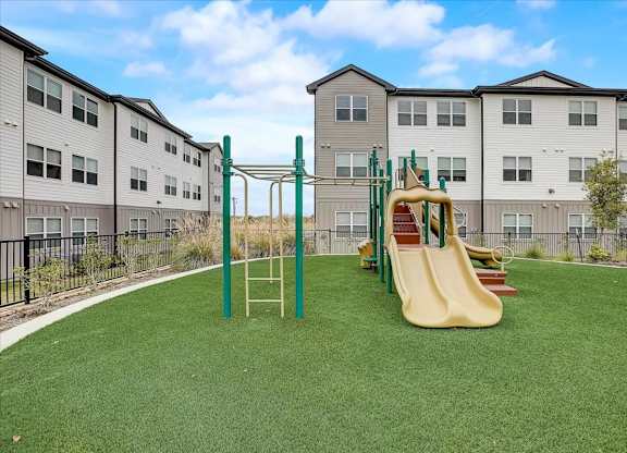 Community playground with slides and climbing features on soft turf, set between apartment buildings.