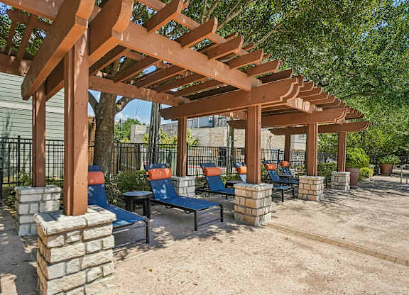 A wooden pergola with blue chairs and a sandy ground.