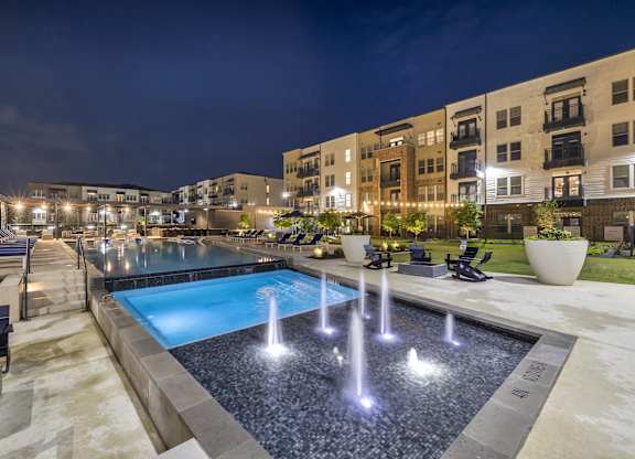 a pool with water fountains in front of an apartment building at night