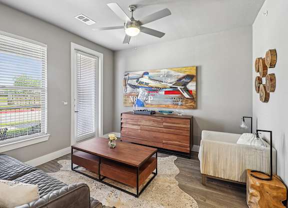 A living room with a grey couch, a coffee table, a painting, a fan, and a window with blinds.