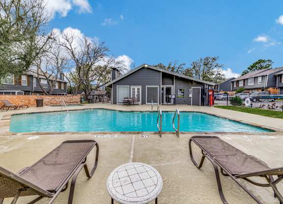 A pool with lounge chairs and a table in the foreground with a building in the background.