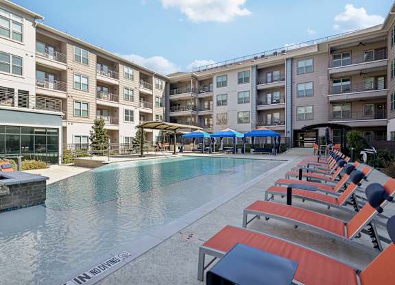 A pool surrounded by sun loungers and umbrellas in front of apartment buildings.