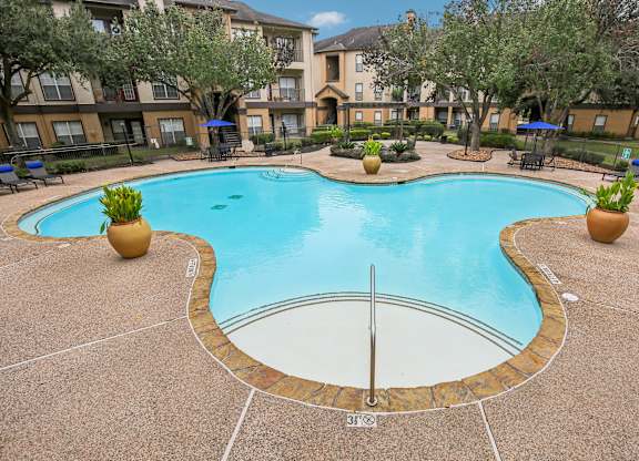 A large outdoor swimming pool surrounded by a concrete patio and a grassy area with trees.