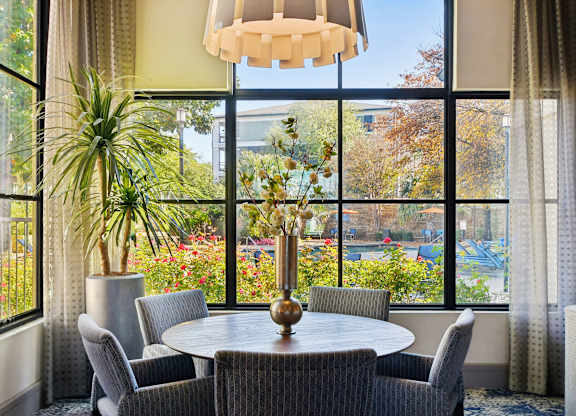 A round table with a vase of flowers on it is surrounded by chairs in a room with large windows.