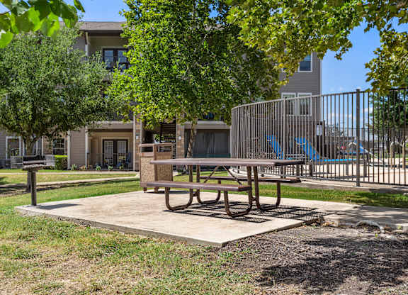 Picnic table and barbecue grill area beside fenced pool and shaded lawn.