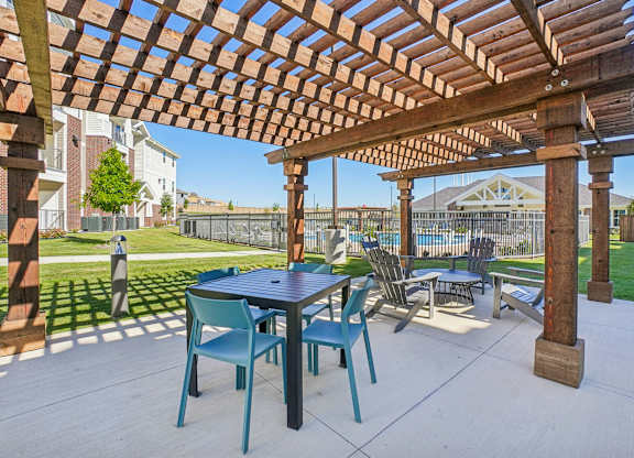 A wooden pergola with a table and chairs is in the foreground of a patio.