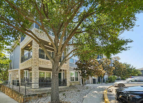 A tree in front of a two-story building with a stone wall and a black car parked on the street.