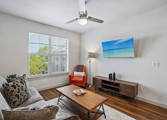 A living room with a grey couch, a wooden coffee table, a red chair, a ceiling fan, and a flat screen TV mounted on the wall.