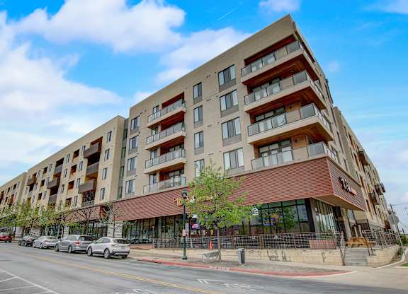 Corner view of mid-rise apartment building with balconies, storefront windows, and street parking.