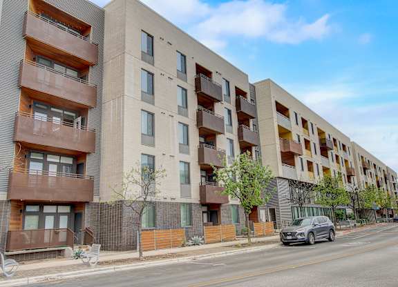 Street-level view of apartment façade with balconies, sidewalks, and parked cars.
