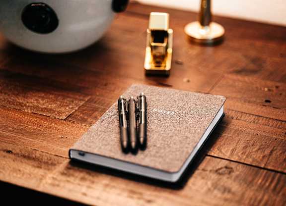 three pens sitting on top of a notebook on a wooden desk