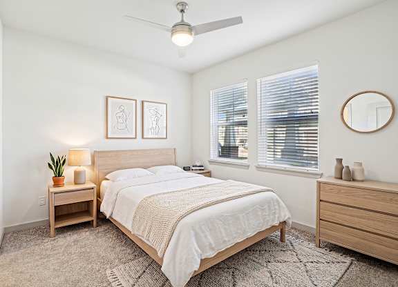 Carpeted bedroom with ceiling fan, two blinded windows, wood nightstand, and dresser with round mirror.
