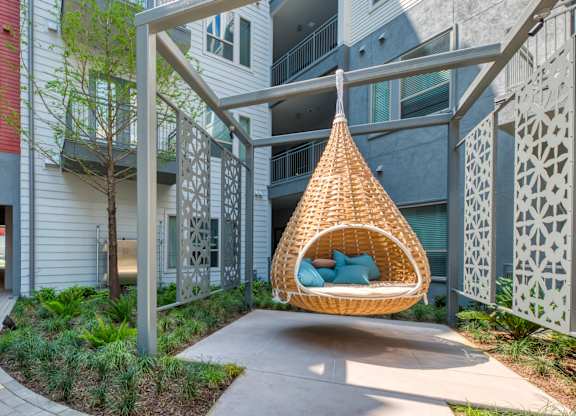 A wicker hanging chair with blue cushions is suspended from a metal frame in a courtyard.