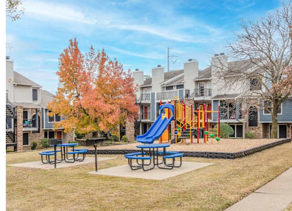 A playground with a blue slide and picnic tables in front of a row of houses.