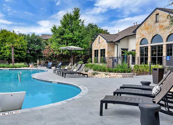 Pool deck with chaise lounges beside a stone-style clubhouse exterior and gardens.
