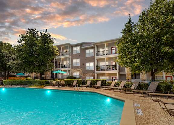 A large swimming pool is surrounded by lounge chairs and trees in front of apartment buildings.